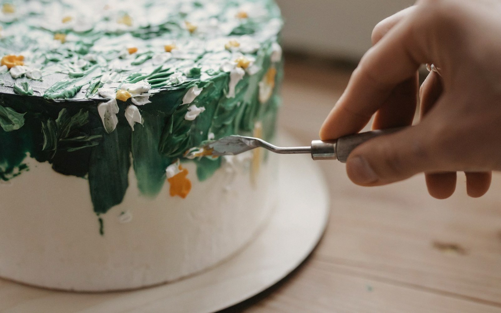 A hand uses a small palette knife to add buttercream flowers along a cake's grassy green border
