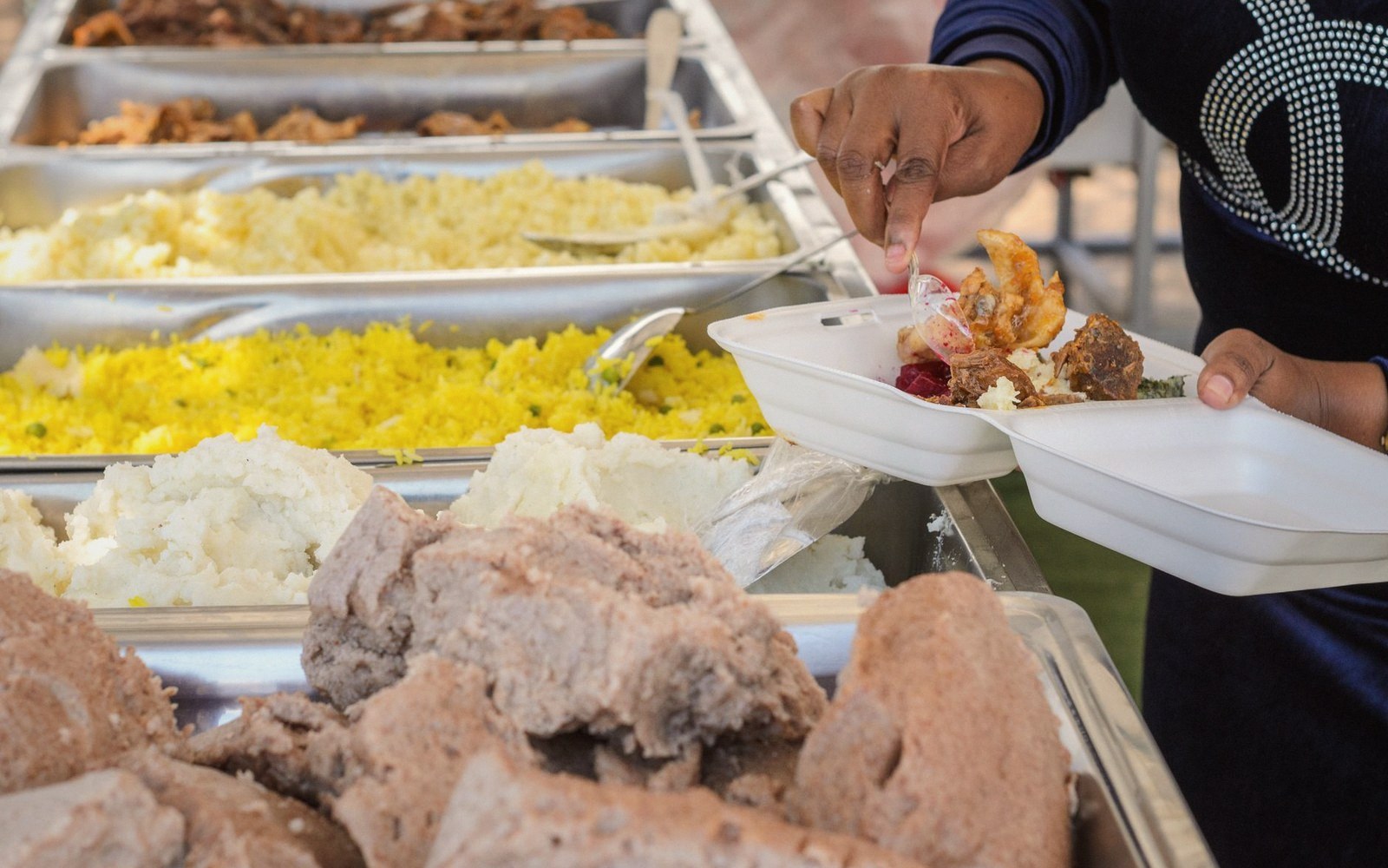 Server trays of rice, mashed potatoes and meat as a hand fills a takeout container
