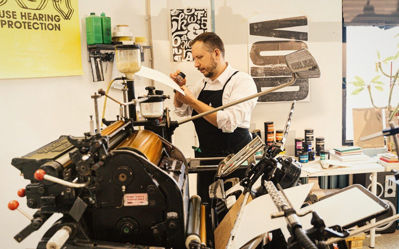 A printer inspects a proof sheet beside an antique letterpress machine in a print studio