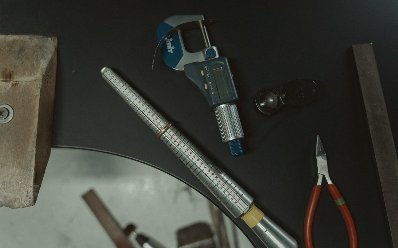 Digital caliper, ring sizer, loupe and pliers arranged on a dark workbench