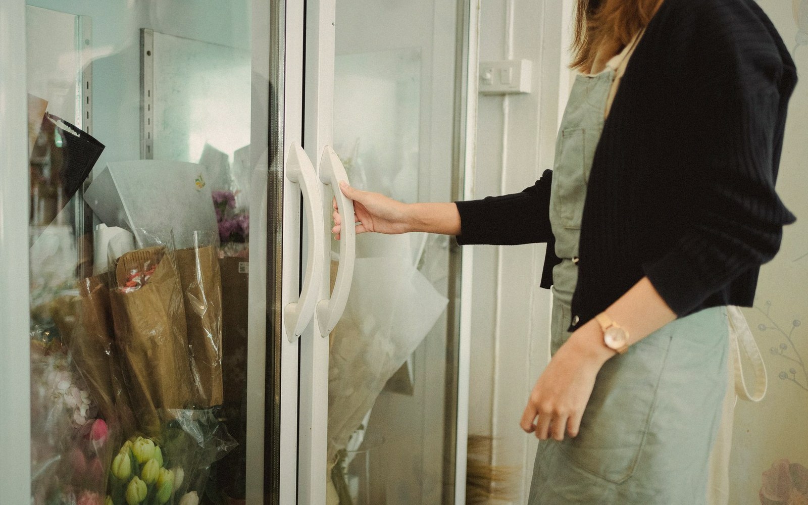 A woman opens a glass cooler door in a flower shop stocked with wrapped bouquets