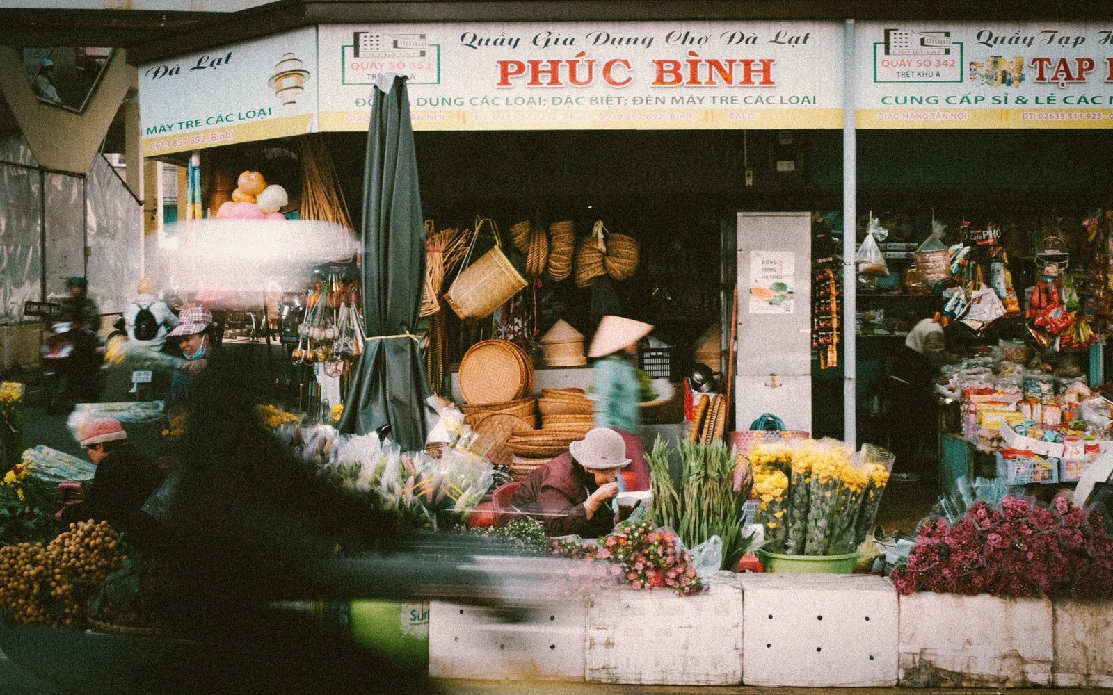 Vendors sell woven baskets and flowers outside a market storefront as a motorbike passes by