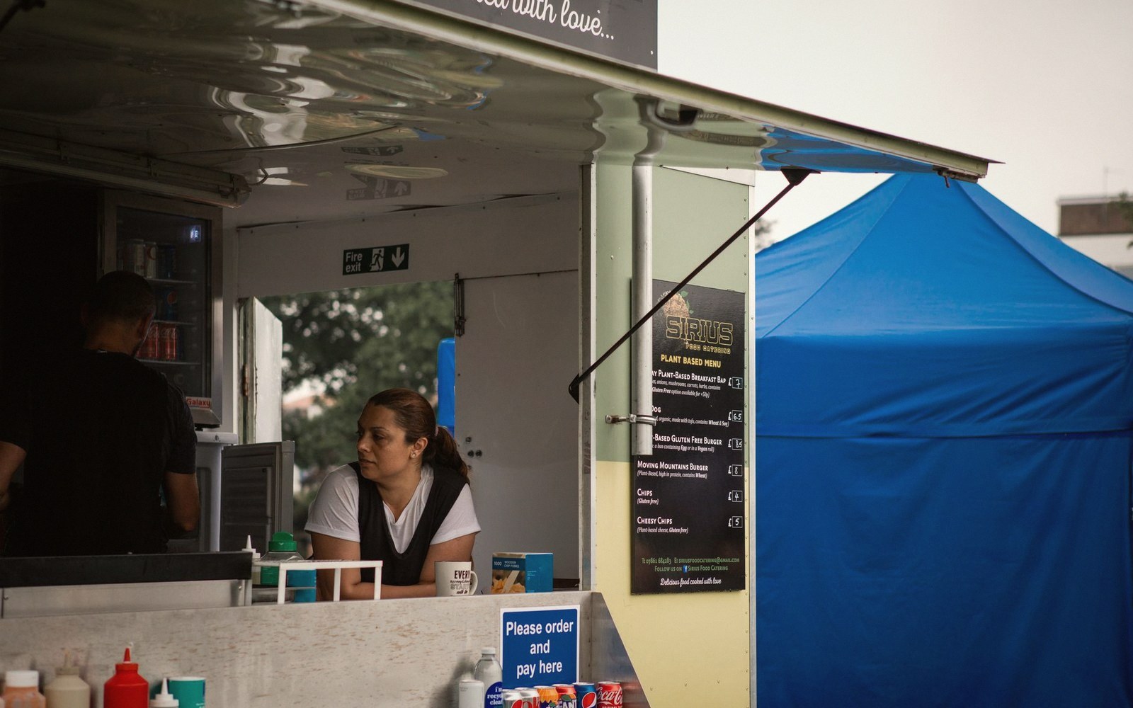 A vendor leans on the counter of a food truck beside a blue pop-up tent