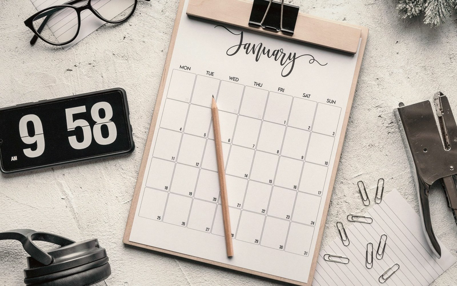 A blank january calendar on a clipboard with a pencil, clock, glasses and stapler nearby