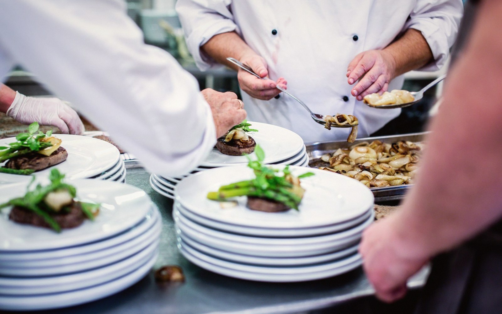 Chefs plate steaks with asparagus and greens on stacked white plates in a kitchen