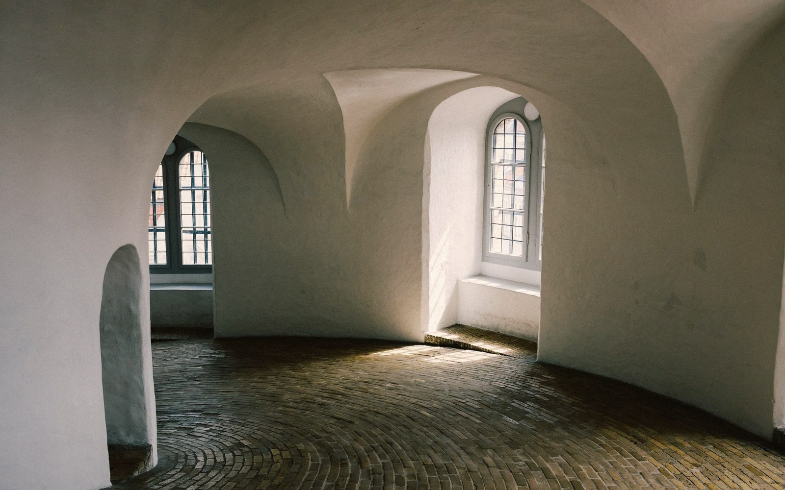 Curved white plaster corridor with arched windows and worn brick flooring